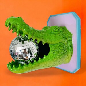 Pode incluir: Escultura de cabe&ccedil;a de jacar&eacute; verde com uma bola de discoteca na boca. A escultura &eacute; montada sobre um fundo roxo claro com uma borda azul, sobre um fundo laranja. Os dentes do jacar&eacute; s&atilde;o vis&iacute;veis.