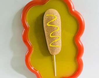 May include: A whimsical, food-themed art piece featuring a corn dog on a stick, adorned with yellow mustard. The corn dog rests on a yellow, glossy surface within a red, wavy-edged frame. The artwork evokes a playful, pop art aesthetic.