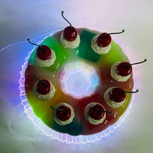 May include: A vibrant, ring-shaped dessert with a rainbow gradient. It's adorned with white cream and bright red cherries with green stems. The dessert is presented on a decorative, scalloped plate with a glowing edge.