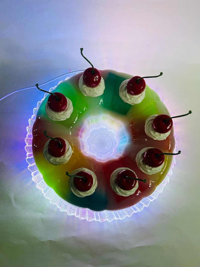 May include: A vibrant, ring-shaped dessert with a rainbow gradient. It's adorned with white cream and bright red cherries with green stems. The dessert is presented on a decorative, scalloped plate with a glowing edge.