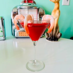 Könnte beinhalten: Ein roter Cocktail in einem Martini-Glas, garniert mit vier Garnelen. Das Glas steht auf einem weißen Tisch vor einem Vintage-Jukebox und einer Keramik-Katzenfigur.