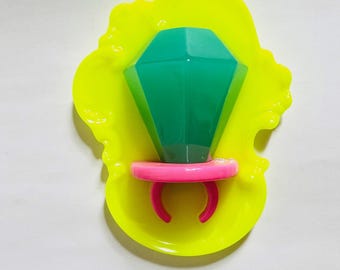May include: A bright green plastic ring with a large diamond-shaped gem on top. The ring is sitting in a yellow plastic mold.