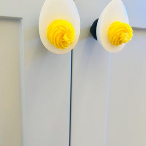 May include: Two white ceramic cabinet knobs shaped like a boiled egg with a yellow yolk. The knobs are mounted on a white cabinet door.