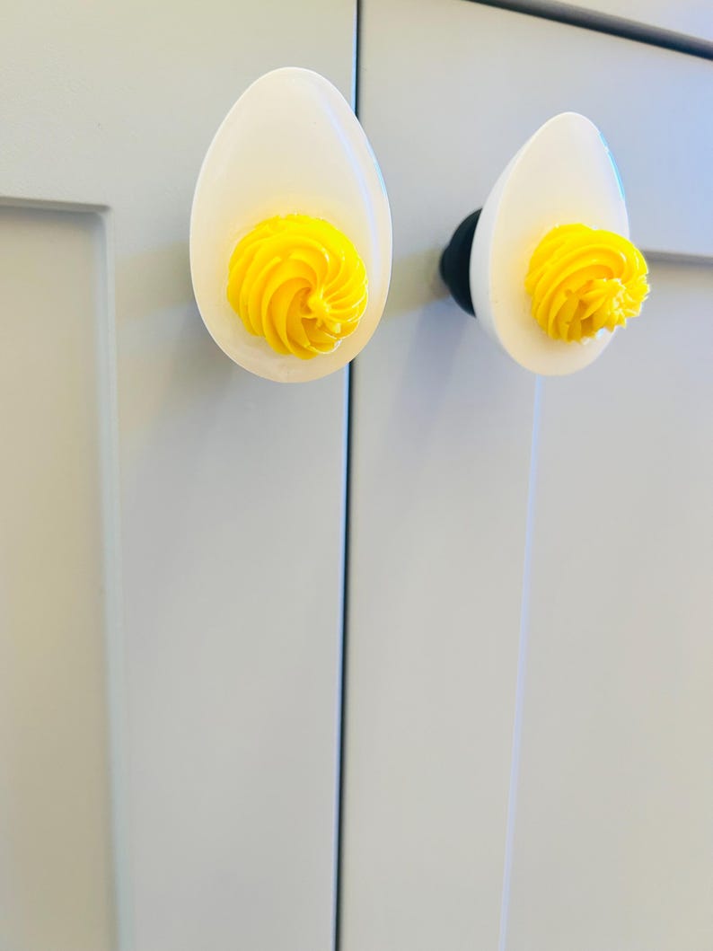 May include: Two white ceramic cabinet knobs shaped like a boiled egg with a yellow yolk. The knobs are mounted on a white cabinet door.