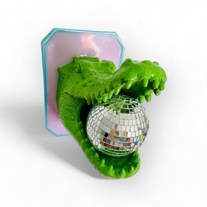Pode incluir: Uma escultura de cabe&ccedil;a de jacar&eacute; verde com a boca aberta segurando uma bola de discoteca. A escultura &eacute; montada em uma placa retangular rosa e azul. O jacar&eacute; tem pele texturizada e dentes afiados.