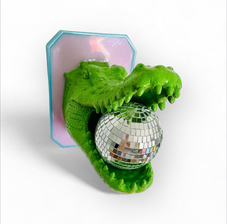 Pode incluir: Uma escultura de cabe&ccedil;a de jacar&eacute; verde com a boca aberta segurando uma bola de discoteca. A escultura &eacute; montada em uma placa retangular rosa e azul. O jacar&eacute; tem pele texturizada e dentes afiados.
