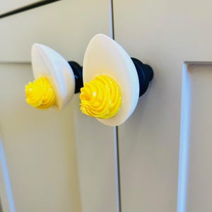 May include: Two white ceramic cabinet knobs shaped like eggs with yellow swirls on the top. The knobs are mounted on a white cabinet door.