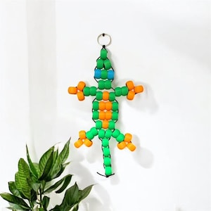 以下が含まれることがあります： 緑、オレンジ、青色のビーズで作られたトカゲの壁掛け。トカゲの上部には吊り下げ用の金属リングが付いています。トカゲの体は緑色のビーズでできており、オレンジと青色のアクセントがあります。白い壁に掛けられています。