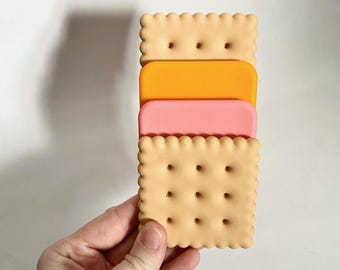 May include: A stack of three silicone cookie cutters shaped like crackers. The top cutter is yellow, the middle cutter is orange, and the bottom cutter is pink.