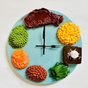 May include: A blue clock with a fork and knife for hands. The clock face is decorated with various food items, including a steak, peas, macaroni and cheese, carrots, baked beans, and green beans.
