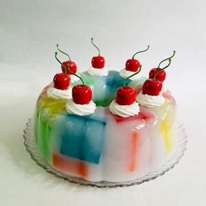 May include: A vibrant, layered jelly dessert in rainbow colours, garnished with whipped cream and bright red cherries. The dessert is presented on a clear, decorative glass plate, creating a festive and appealing look.