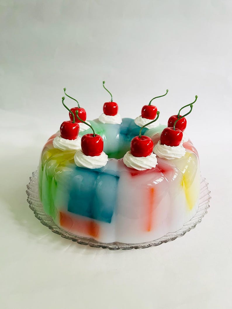May include: A vibrant, layered jelly dessert in rainbow colours, garnished with whipped cream and bright red cherries. The dessert is presented on a clear, decorative glass plate, creating a festive and appealing look.