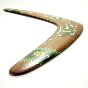 Op de afbeelding: Een houten boemerang met een gebogen vorm, beschilderd met bruine, groene, gele en witte ontwerpen. De boemerang heeft een glanzende afwerking en staat tegen een witte achtergrond. Het ontwerp omvat bloemen en geometrische patronen.