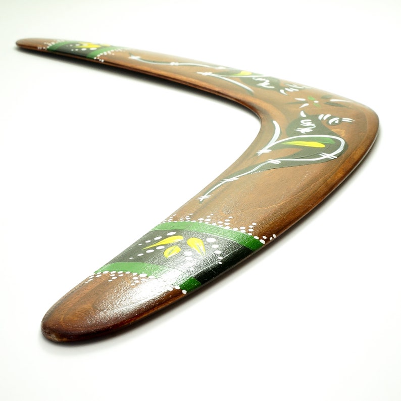 Op de afbeelding: Een houten boemerang met een gebogen vorm, beschilderd met bruine, groene, gele en witte ontwerpen. De boemerang heeft een glanzende afwerking en staat tegen een witte achtergrond. Het ontwerp omvat bloemen en geometrische patronen.