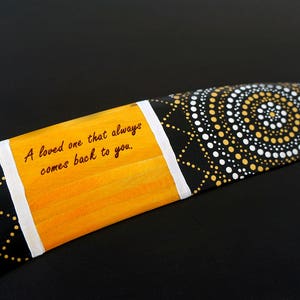 K&ouml;nnte beinhalten: Ein Bumerang, der mit schwarzen, wei&szlig;en und goldenen Punkten bemalt ist. Der Bumerang hat einen gelben Streifen mit dem Text "A loved one that always comes back to you."
