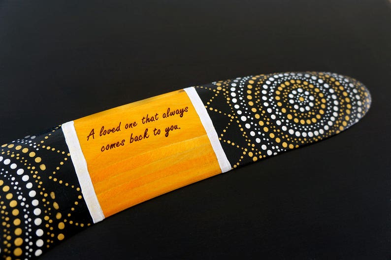 K&ouml;nnte beinhalten: Ein Bumerang, der mit schwarzen, wei&szlig;en und goldenen Punkten bemalt ist. Der Bumerang hat einen gelben Streifen mit dem Text "A loved one that always comes back to you."