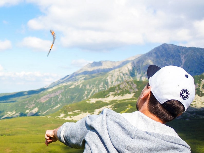 K&ouml;nnte beinhalten: Eine Person mit einer wei&szlig;en Baseballkappe mit schwarzem Logo wirft einen Bumerang in einer Berglandschaft.
