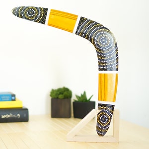 K&ouml;nnte beinhalten: Ein Holz-Bumerang mit einem Muster aus schwarzen, wei&szlig;en und gelben Punkten. Der Bumerang steht auf einem Holzst&auml;nder.