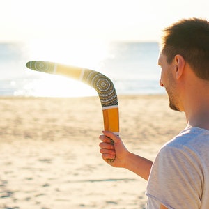 K&ouml;nnte beinhalten: Eine Person wirft einen Bumerang mit einem farbenfrohen Aborigine-Design an einem Sandstrand. Der Bumerang ist aus Holz und hat ein gelbes, schwarzes und wei&szlig;es Muster.