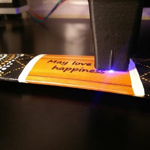 K&ouml;nnte beinhalten: Ein Lasergravierer graviert die Worte "May love happiness" auf eine Holzoberfl&auml;che mit einem schwarzen, wei&szlig;en und goldenen Muster.