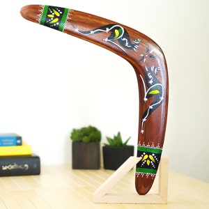 Op de afbeelding: Een houten boemerang met een gebogen ontwerp, met een kangoeroe-illustratie in zwart, wit en geel. De boemerang heeft groene en gele accenten aan de uiteinden en wordt tentoongesteld op een houten standaard.