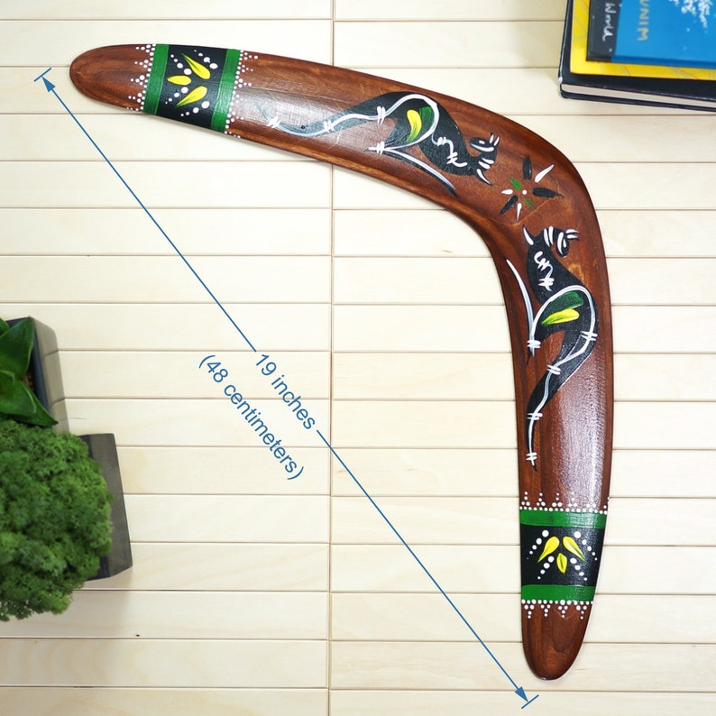 Op de afbeelding: Een houten boemerang, 48 centimeter lang, met een donkerbruine afwerking. Het heeft handgeschilderde ontwerpen in zwart, wit en geel, waaronder kangoeroe-figuren en geometrische patronen. De boemerang heeft groene accenten en witte gestippelde randen.