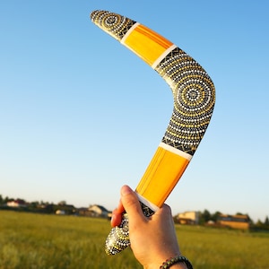 K&ouml;nnte beinhalten: Ein gelbes und schwarzes Bumerang mit einem komplizierten Punktmuster. Das Bumerang wird in einer Hand vor einem blauen Himmel gehalten.