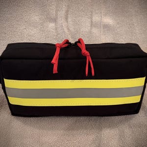 May include: Black rectangular bag with a yellow and gray reflective stripe design. The bag has two red zipper pulls and small black handles on each side. The bag is sitting on a gray textured surface.