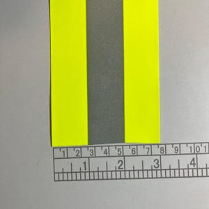 May include: A reflective safety strip with a gray center and bright yellow sides. The strip is measured with a ruler, showing approximately 4 inches in length. The material appears to be durable and suitable for visibility.