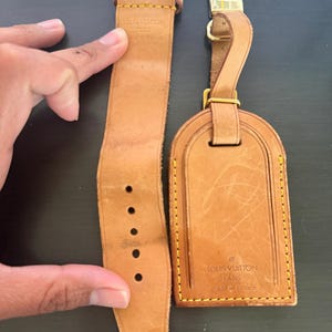 Puede incluir: Un juego de correa de reloj y etiqueta de equipaje de cuero marrón claro. La correa del reloj tiene varios agujeros y una hebilla dorada. La etiqueta de equipaje tiene un cierre dorado y el texto "LOUIS VUITTON PARIS made in France".