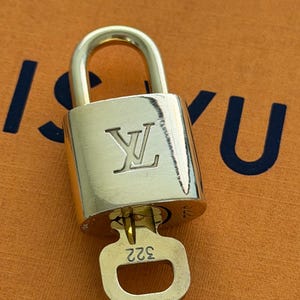 Op de afbeelding: Een goudkleurig hangslot met bijpassende sleutel. Het hangslot heeft het reliëf "LV" logo en de sleutel is gemarkeerd met het nummer "322". Een klassiek accessoire.