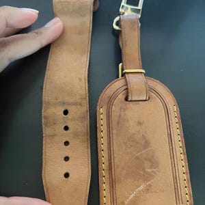 Puede incluir: Un juego de correa de reloj y etiqueta de equipaje de cuero marrón claro. La correa del reloj tiene una hebilla y varios orificios de ajuste. La etiqueta de equipaje es rectangular con bordes redondeados, costuras doradas y el logotipo "LOUIS VUITTON".