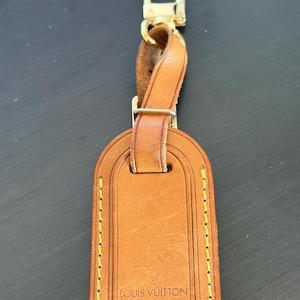May include: A brown leather luggage tag with gold-colored hardware. The tag has a rectangular shape with rounded top edges and yellow stitching. The text "LOUIS VUITTON PARIS made in France" is printed on the tag.