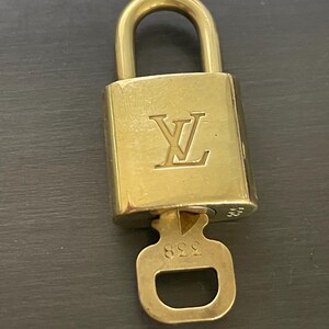 Op de afbeelding: Een goudkleurig hangslot met bijpassende sleutel. Het hangslot heeft het reliëf "LV" logo op de voorkant. De sleutel zit in het slot, met het nummer "338" zichtbaar op de sleutel.