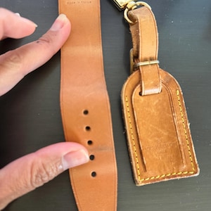 Puede incluir: Correa de reloj y etiqueta de equipaje de cuero color canela con herrajes dorados. La correa del reloj tiene cinco agujeros y el texto "LOUIS VUITTON PARIS made in france". La etiqueta de equipaje tiene el mismo texto y costuras amarillas.