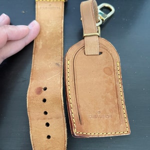 Puede incluir: Una correa de reloj y una etiqueta de equipaje de cuero marrón claro. La correa del reloj tiene varios agujeros de ajuste y la marca "LOUIS VUITTON" es visible. La etiqueta de equipaje tiene un cierre dorado y la marca "LOUIS VUITTON" está grabada.