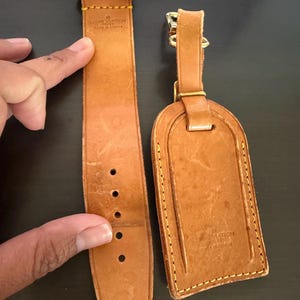Puede incluir: Una etiqueta de equipaje y una correa de cuero marrón claro con herrajes dorados. La correa tiene varios orificios de ajuste. La etiqueta y la correa están estampadas con "LOUIS VUITTON made in France". Los artículos están sobre una superficie oscura.