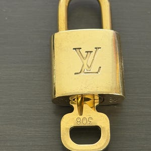 May include: A gold-colored padlock with a matching key. The padlock features the embossed "LV" logo on the front. The key has the number "308" engraved on it. The lock and key are set against a dark gray background.
