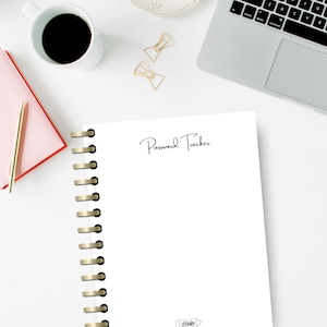 Könnte beinhalten: Ein weißes Notizbuch mit der Aufschrift "Password Tracker" in Schreibschrift. Das Notizbuch hat eine goldfarbene Spiralbindung und ist von Büroartikeln umgeben, darunter ein Laptop, eine Kaffeetasse und Büroklammern.