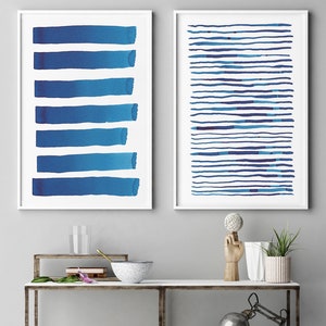 May include: Two framed abstract art prints. One features six horizontal blue brushstrokes, the other has numerous blue horizontal lines. The prints are displayed above a shelf with decorative items.