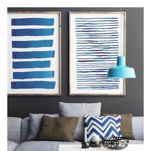 May include: Two framed abstract art prints with blue brushstrokes hang above a gray sofa with brown and blue pillows. A turquoise pendant lamp hangs from the ceiling. A white coffee table and a gray ottoman complete the living room setting.
