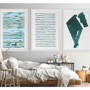 May include: Three framed abstract art prints. The prints feature watercolor designs in shades of teal and blue. One print has horizontal lines, another has diagonal dashes, and the third has geometric shapes. The prints are displayed above a bed with white bedding.