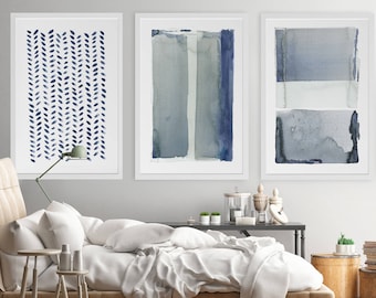 May include: Three framed abstract watercolor paintings in shades of blue and gray. The artwork features various geometric and organic shapes, including vertical stripes and leaf-like patterns. The paintings are displayed above a bed with white bedding.