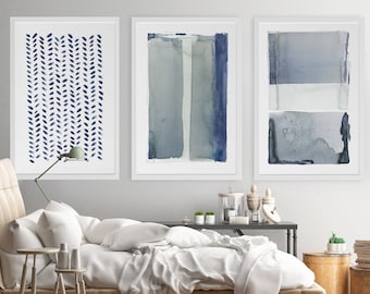 May include: Three framed abstract watercolor prints in shades of blue and gray. The artwork features vertical stripes, leaf-like patterns, and layered rectangular shapes. The prints are displayed above a bed with white bedding.