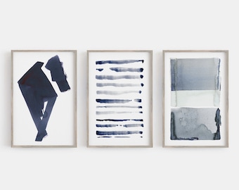 May include: Three framed abstract watercolor prints. The prints feature navy and gray watercolor designs. One print has a geometric shape, another has horizontal lines, and the third has layered rectangles. The frames are a light wood color.