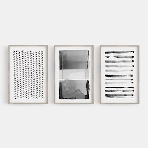 May include: Three framed abstract art prints. The prints feature black and gray watercolor designs. One print has a pattern of small, vertical leaf shapes. Another has horizontal bands of color. The third has a series of horizontal lines.