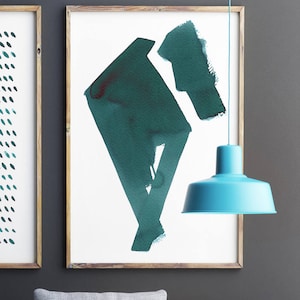 May include: Two framed abstract art prints with teal and dark teal watercolor designs. A light blue pendant lamp hangs in front of the artwork. The frames are made of light brown wood. The background is a dark gray wall.