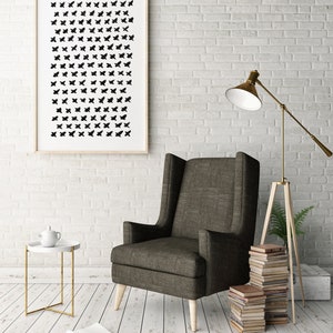 May include: A framed art print with a pattern of black shapes hangs on a white brick wall. A dark gray armchair with light wood legs sits next to a small white side table with a gold frame. A floor lamp and a stack of books are also visible.