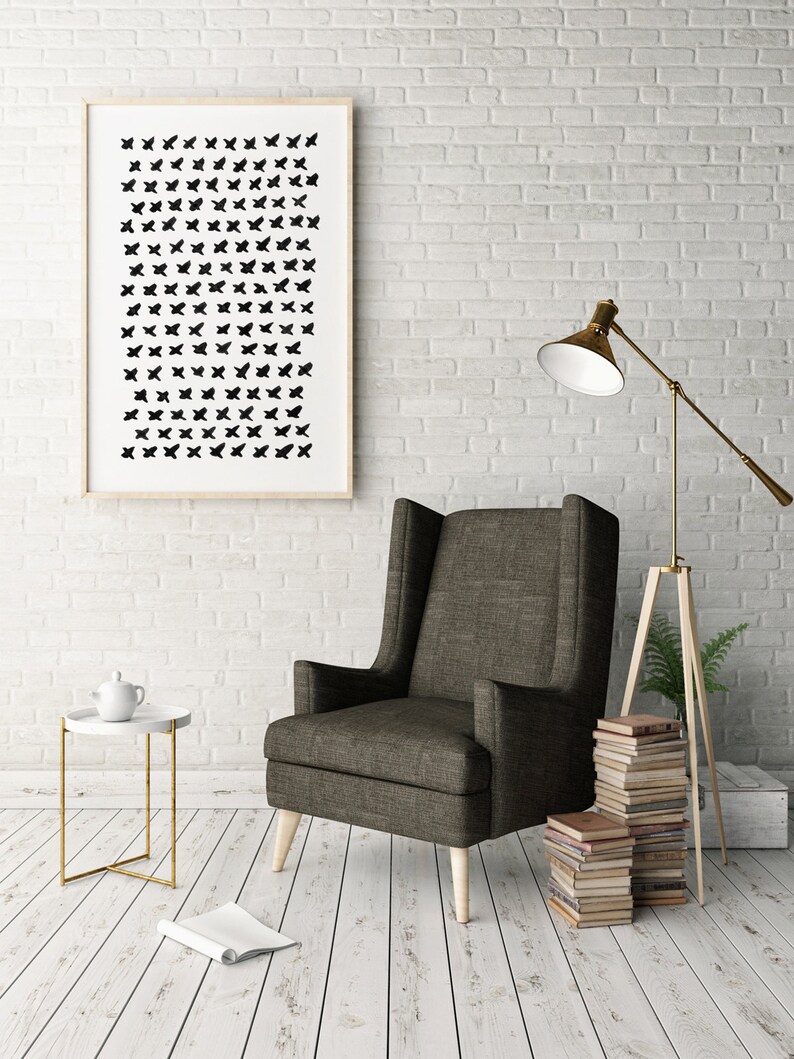 May include: A framed art print with a pattern of black shapes hangs on a white brick wall. A dark gray armchair with light wood legs sits next to a small white side table with a gold frame. A floor lamp and a stack of books are also visible.