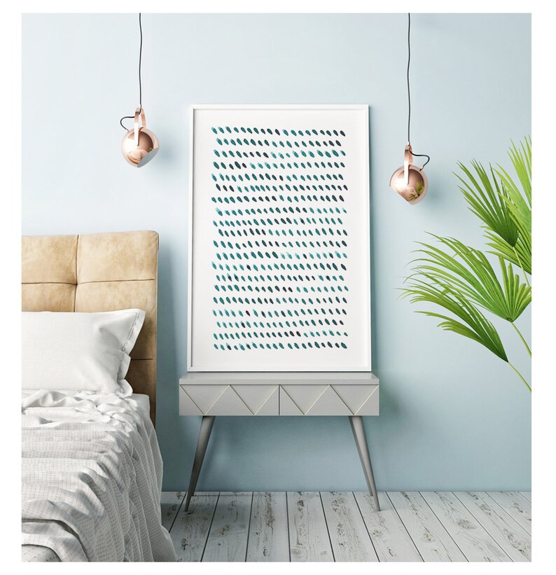May include: A framed abstract art print featuring a pattern of teal and dark blue brushstrokes on a white background. The artwork is displayed on a gray nightstand in a bedroom setting. The room has light blue walls and a bed with a beige headboard.
